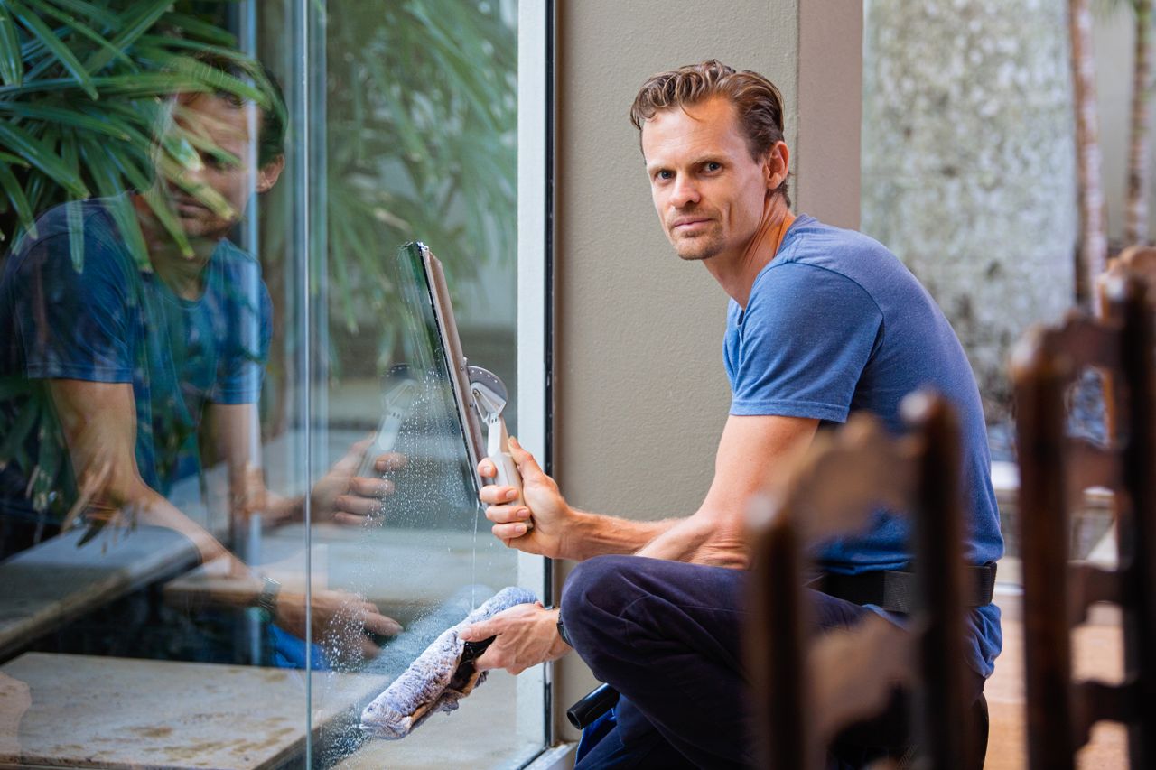 gallery-01 A man wearing a blue t-shirt is cleaning a large glass pane indoors with a squeegee.